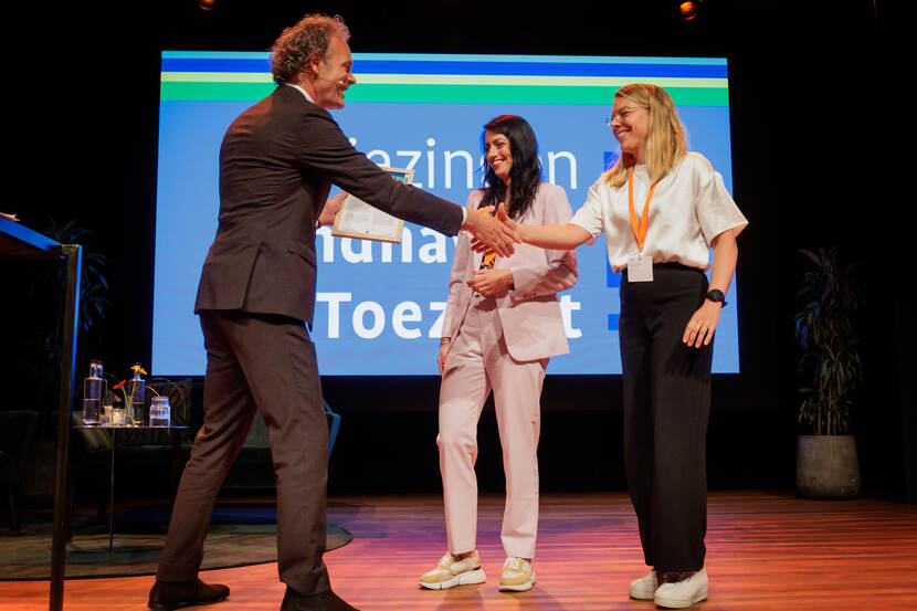 Man schudt hand met vrouw op podium, tweede vrouw kijkt toe