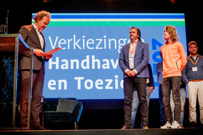 Man in pak leest op een podium rapport voor, man en jongen luisteren