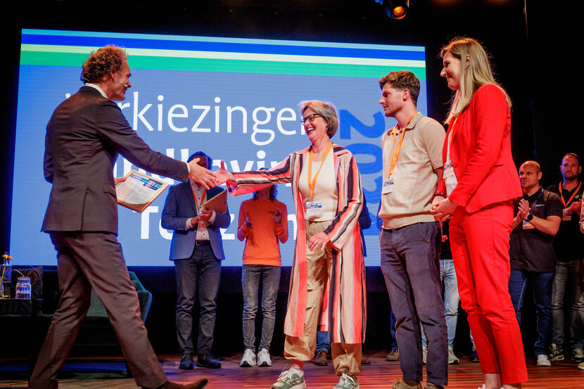 Man in pak schudt hand van vrouw op podium, man en vrouw kijken toe