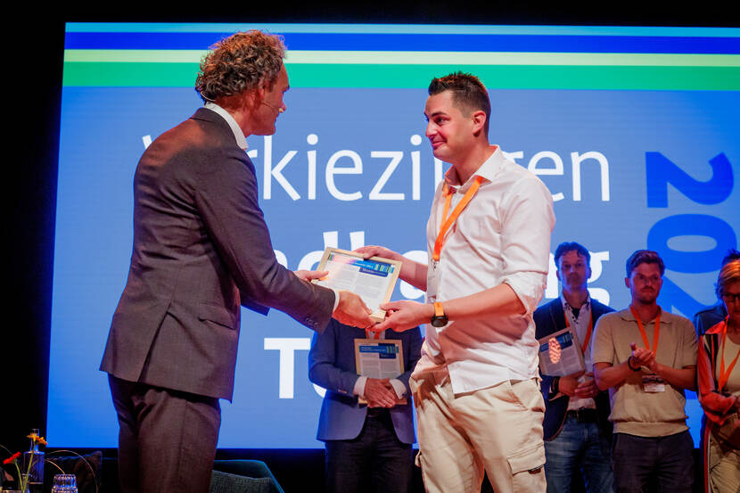 Man in pak overhandigt oorkonde op een podium aan andere man